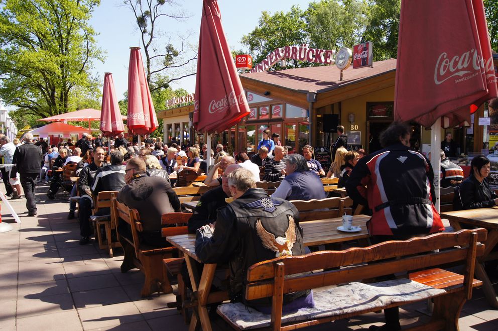 Gäste und Biker bei Essen und Getränken unter roten Schirmen im Biergarten der Spinner-Brücke