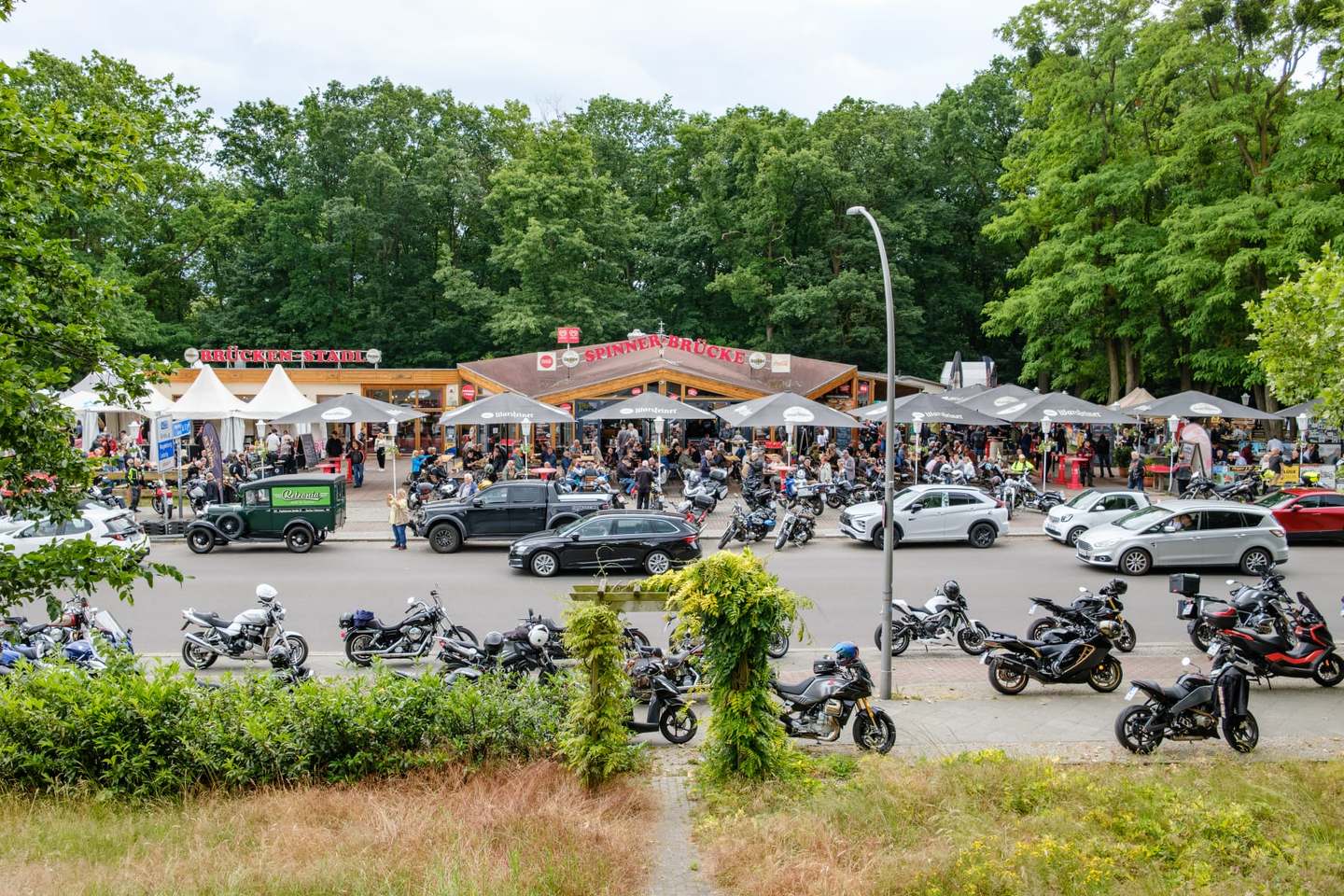 Die Spinner-Brücke von der gegenüberliegenden Straßenseite: Biergarten voller Gäste unter schwarzen Sonnenschirmen, davor Reihen abgestellter Motorräder, dahinter das Gasthaus mit Giebeldach und der Grunewald.